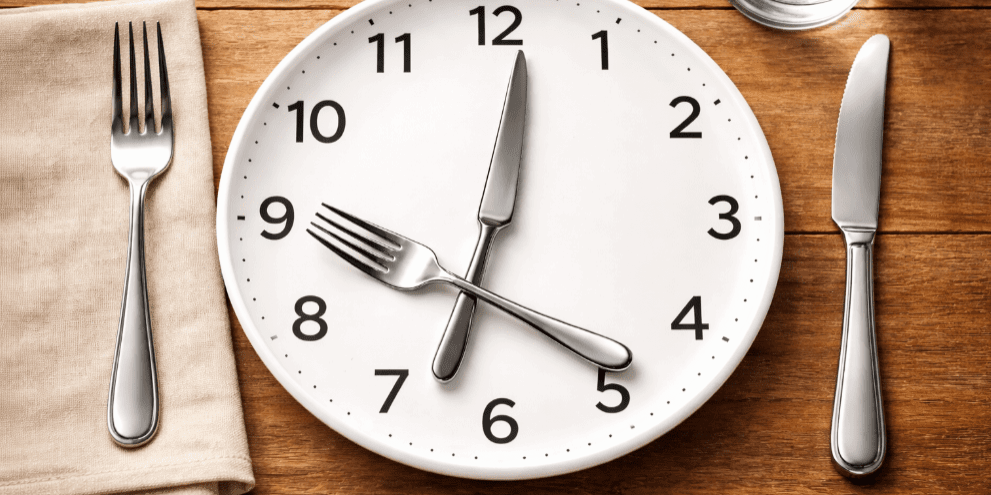 White dinner plate designed as a clock with a fork and knife as clock hands on a wooden table, symbolizing intermittent fasting and meal timing.